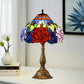 Stained glass table lamp with red rose shade and bronze base on a tabletop.